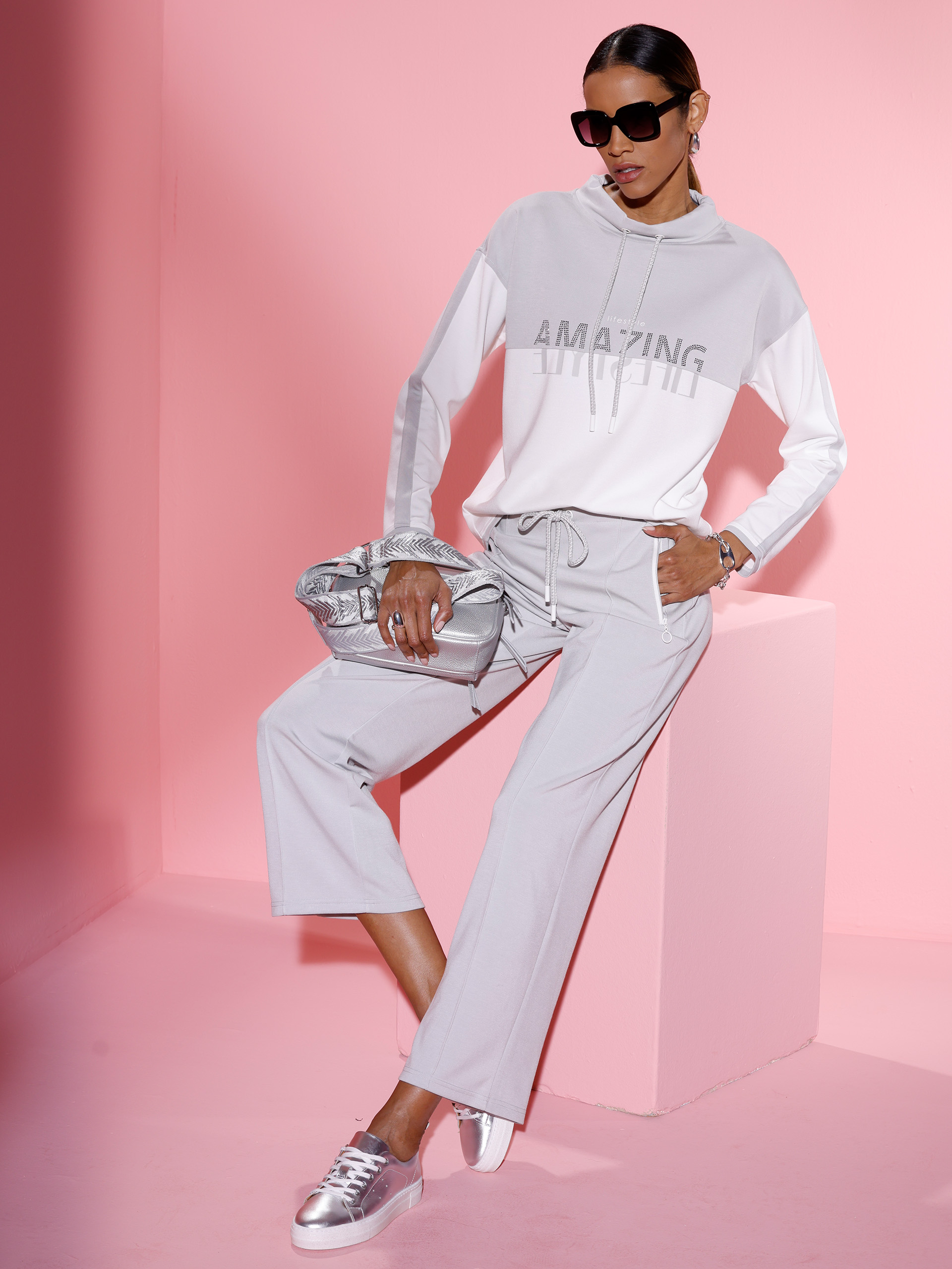 Ein Model sitzt vor rosa Hintergrund, trägt einen hellgrauen Hoodie, weite Hose, silberne Tasche und silberfarbene Sneaker.