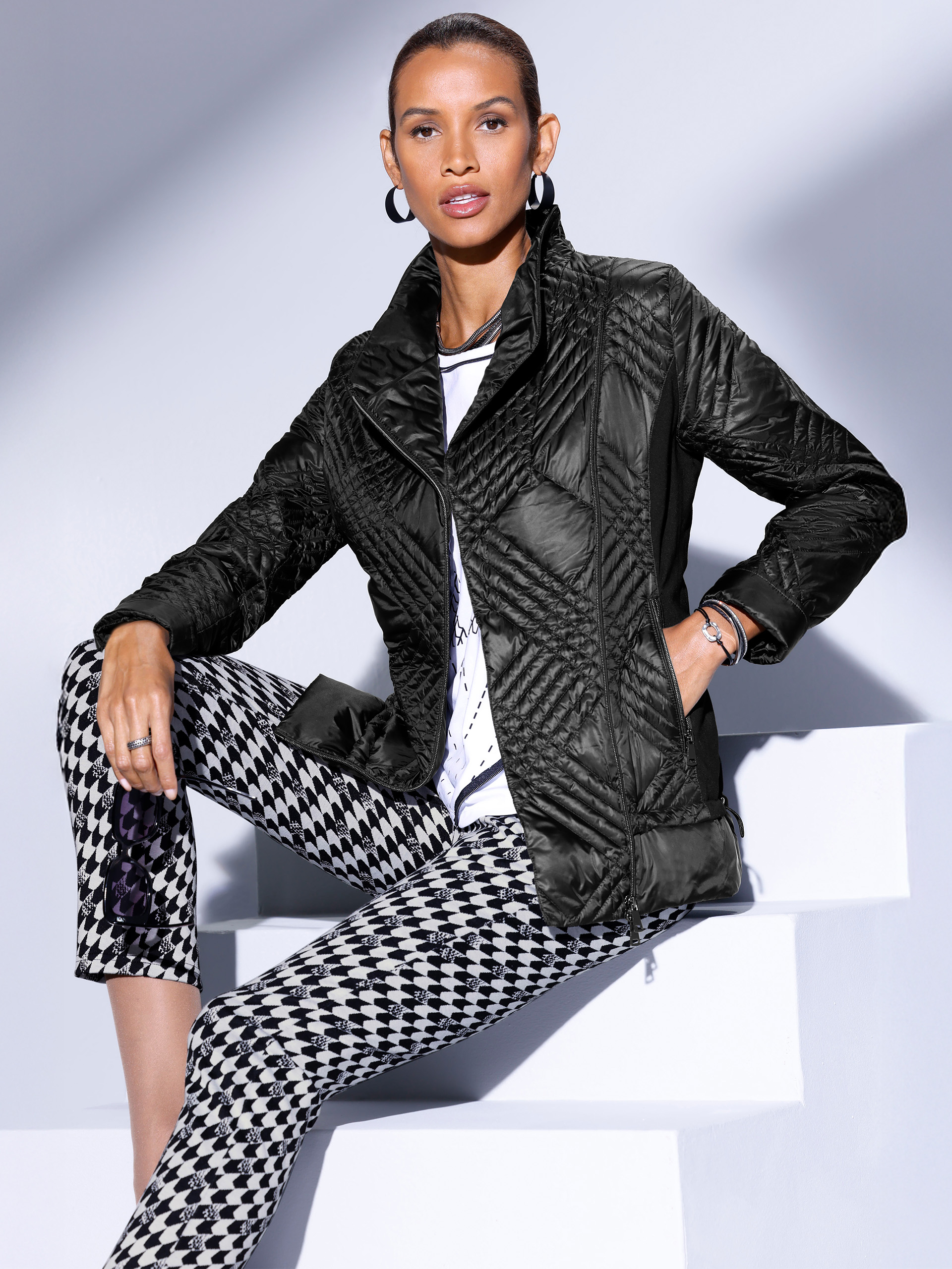 Ein Model trägt eine schwarze, gesteppte Jacke mit einem modernen Schnitt und enganliegende Hosen mit einem schwarz-weißen Hahnentrittmuster. Der Hintergrund ist hellgrau.