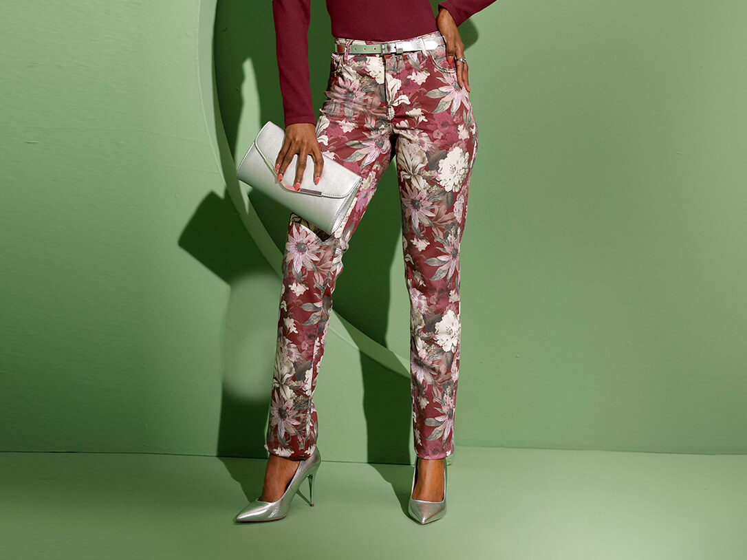 Ein Model trägt eine bordeauxfarbene Bluse und gemusterte Hosen mit Blumenmotiven vor einer grünen Wand.