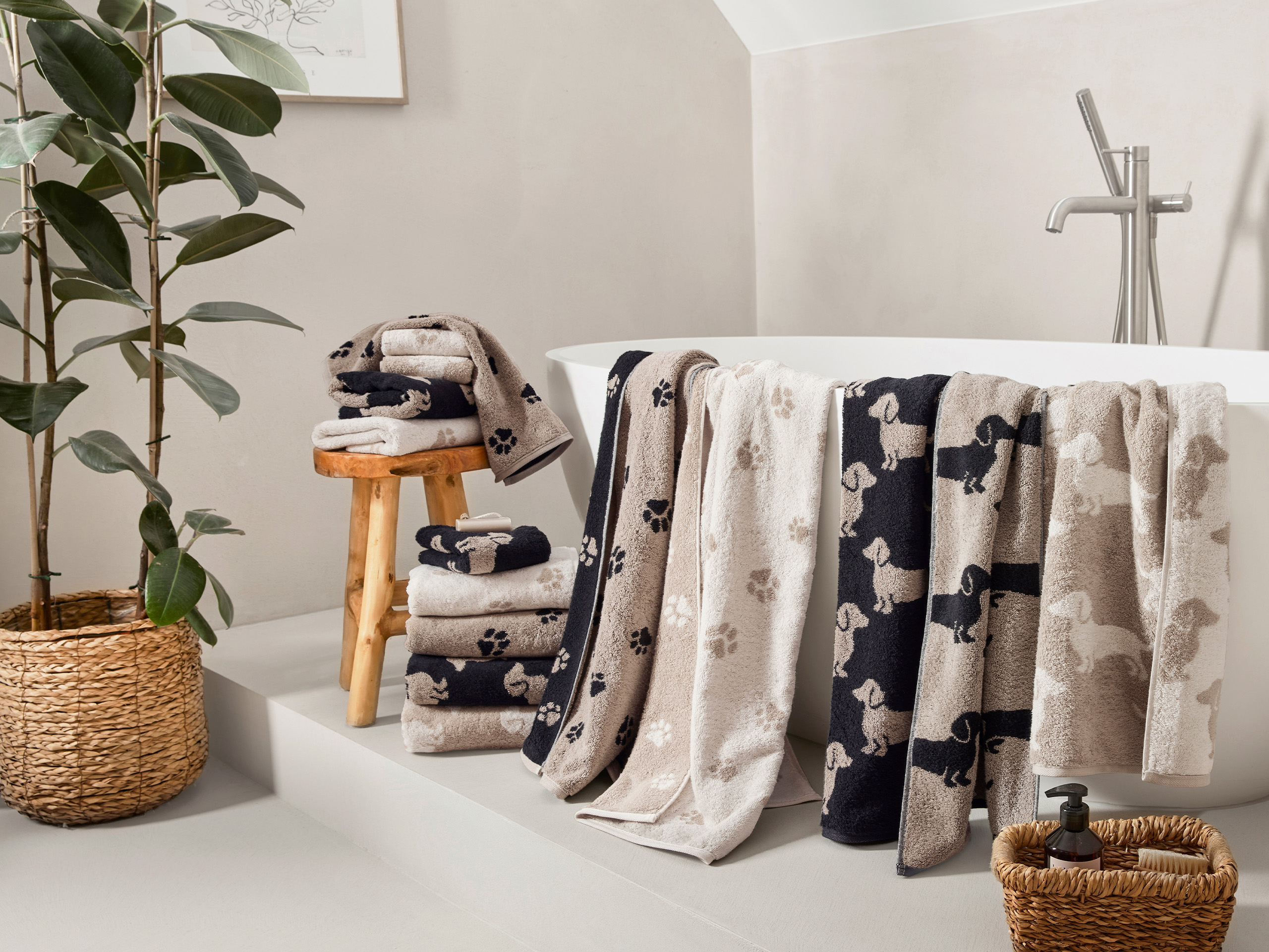 Verschiedene Handtücher mit Hunde-Motiven im Badezimmer, neben Pflanze und Wanne.
