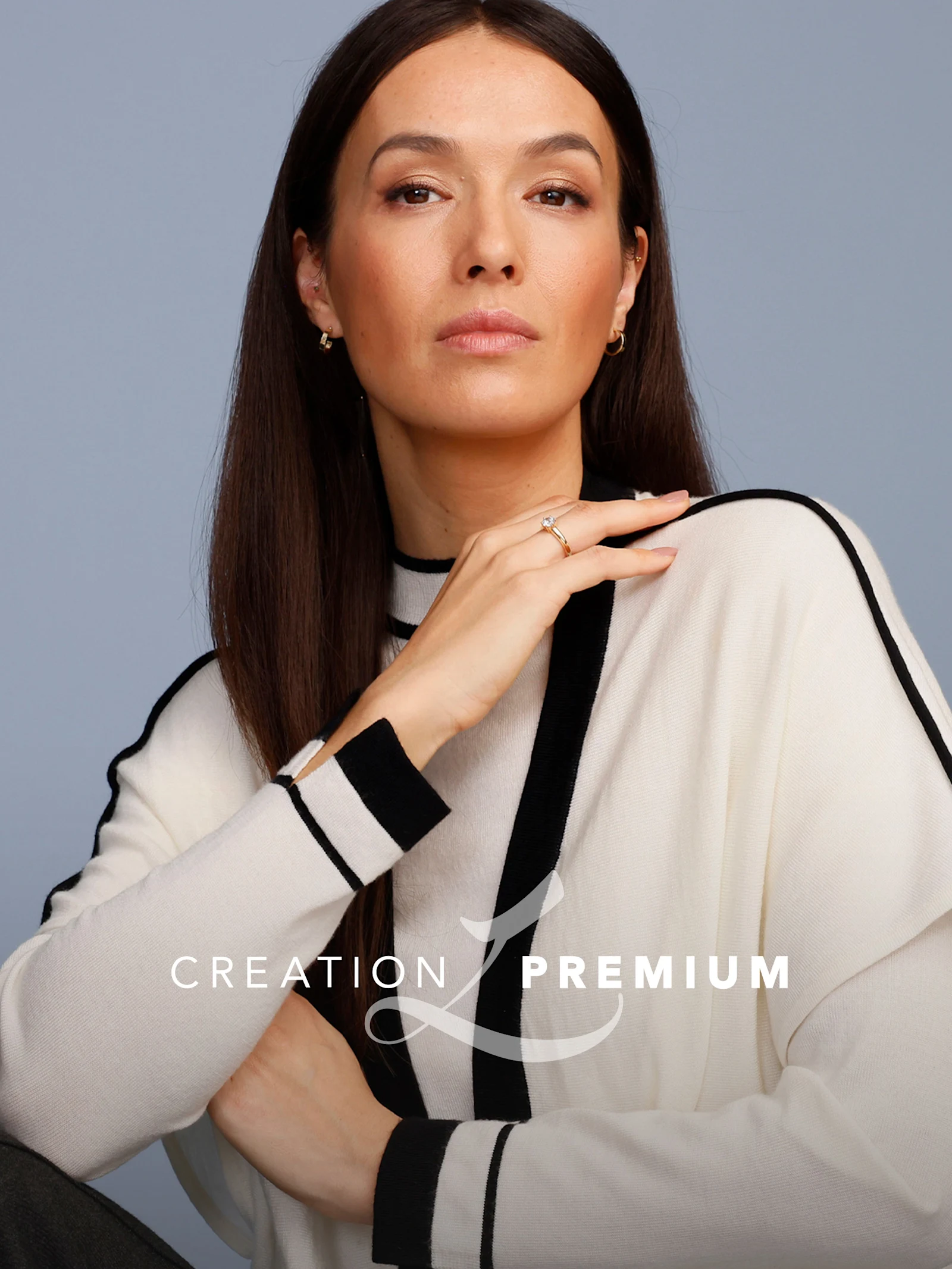 Ein Model trägt einen cremefarbenen Pullover mit schwarzen Streifen, sitzt vor graublauem Hintergrund. Text: 'Creation Premium'.