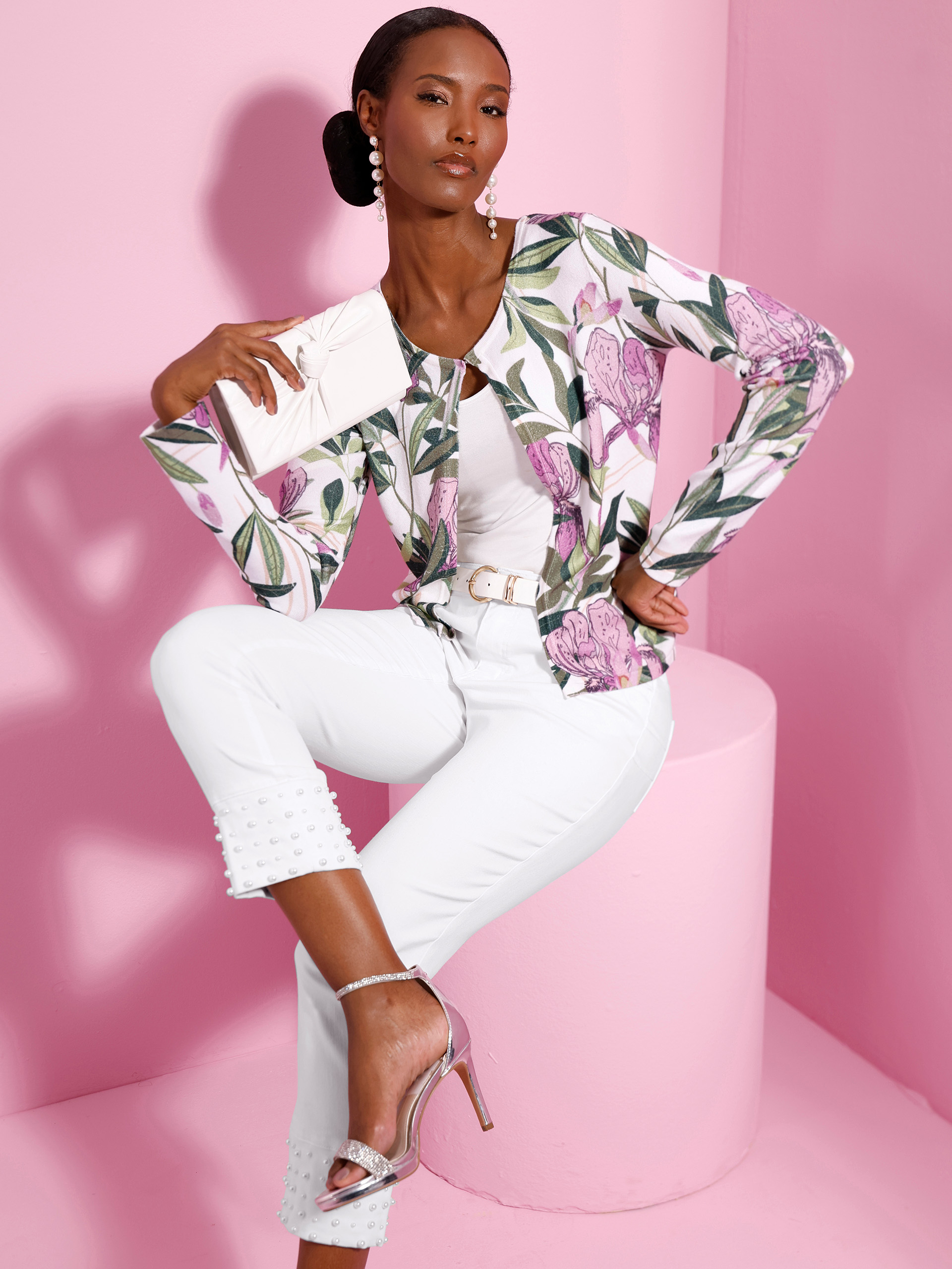 Ein Model sitzt vor rosa Hintergrund, trägt eine weiße Hose mit Perlenbesatz, ein florales Oberteil und silberne High Heels, hält eine helle Clutch.