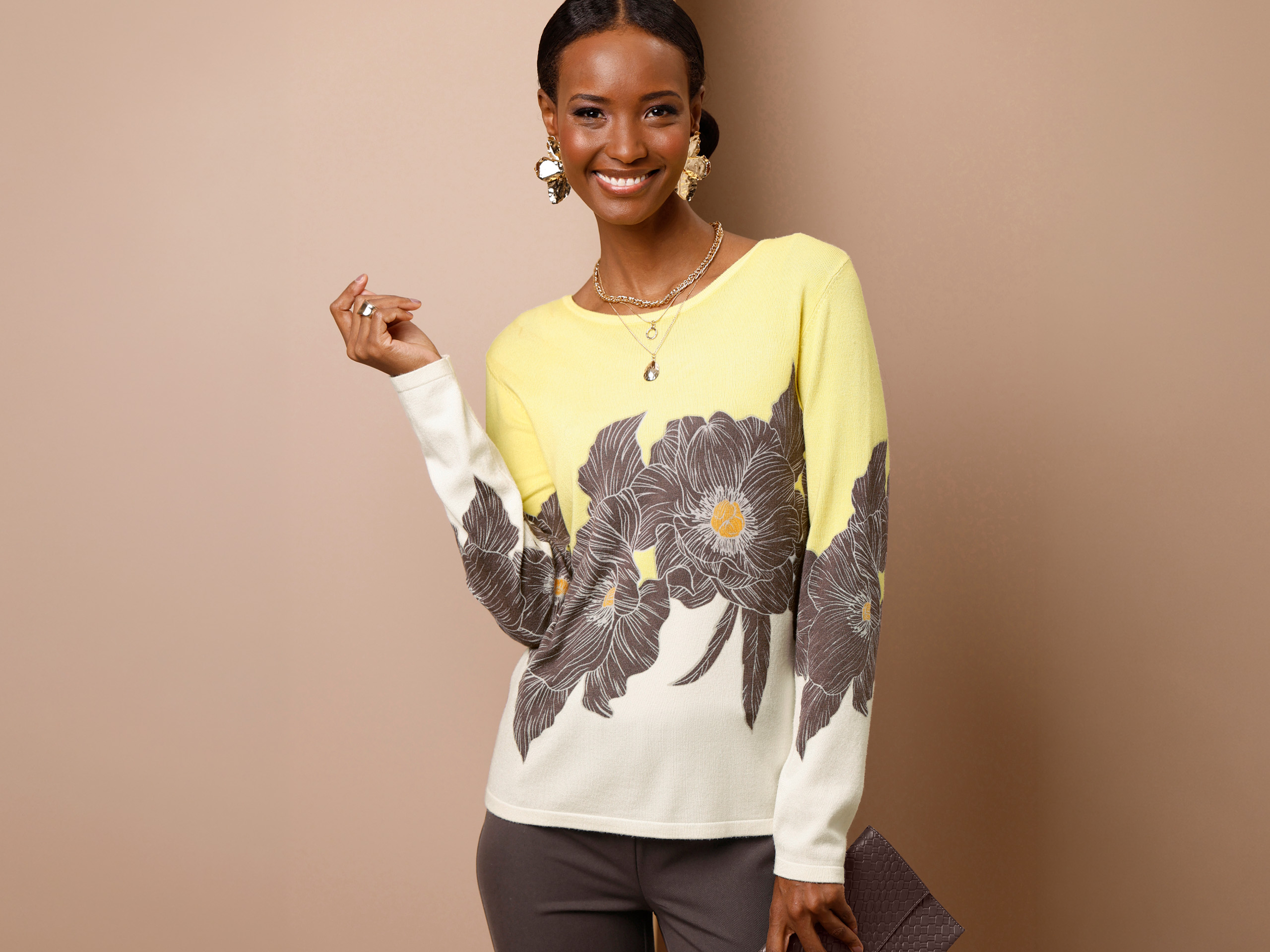 Ein Model trägt einen gelb-weißen Pullover mit großem Blumenmuster und eine dunkle Hose. Sie hält eine Clutch und lächelt. Der Hintergrund ist beige.