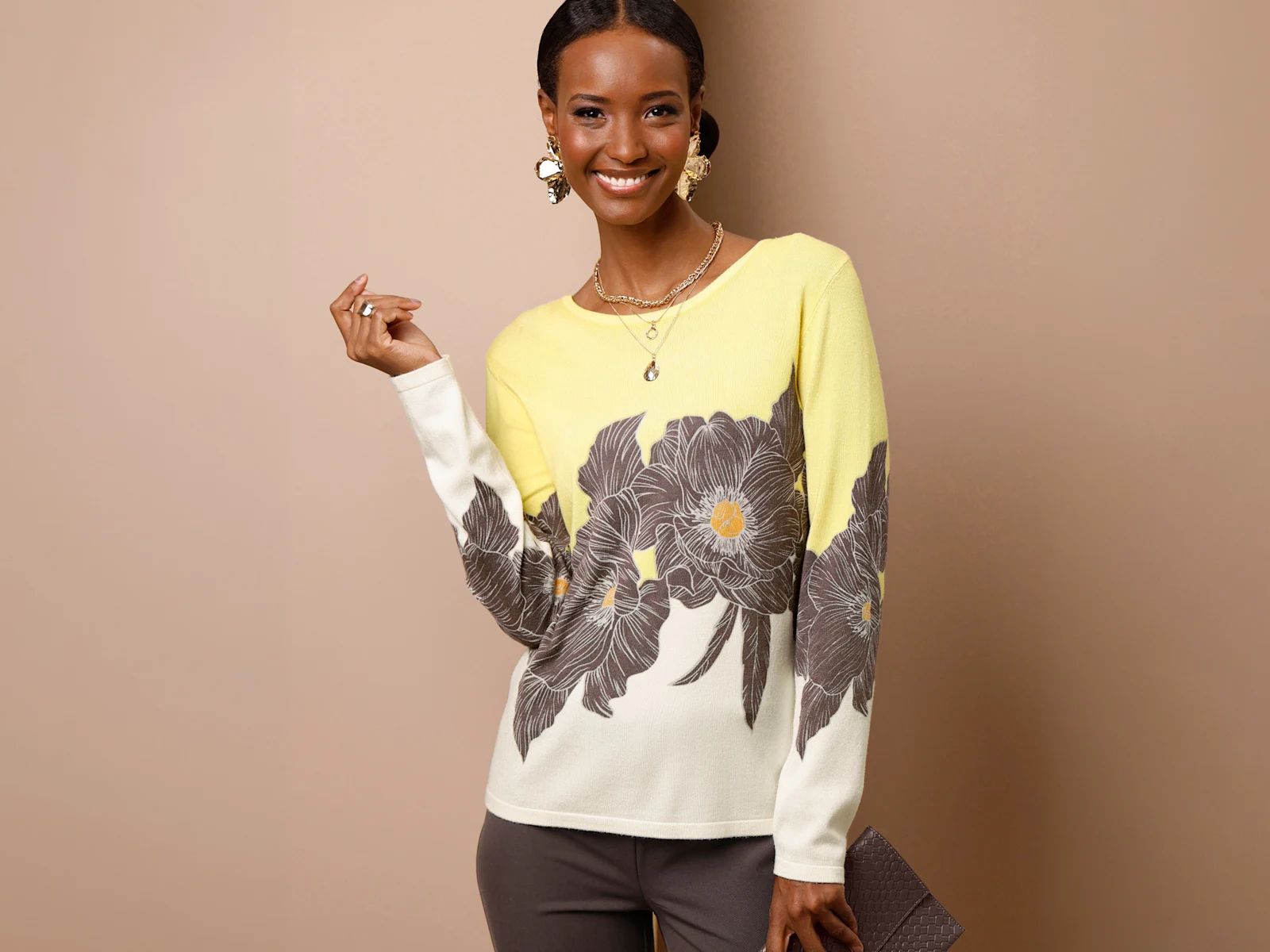 Ein Model trägt einen gelb-weißen Pullover mit großem Blumenmuster und eine dunkle Hose. Sie hält eine Clutch und lächelt. Der Hintergrund ist beige.