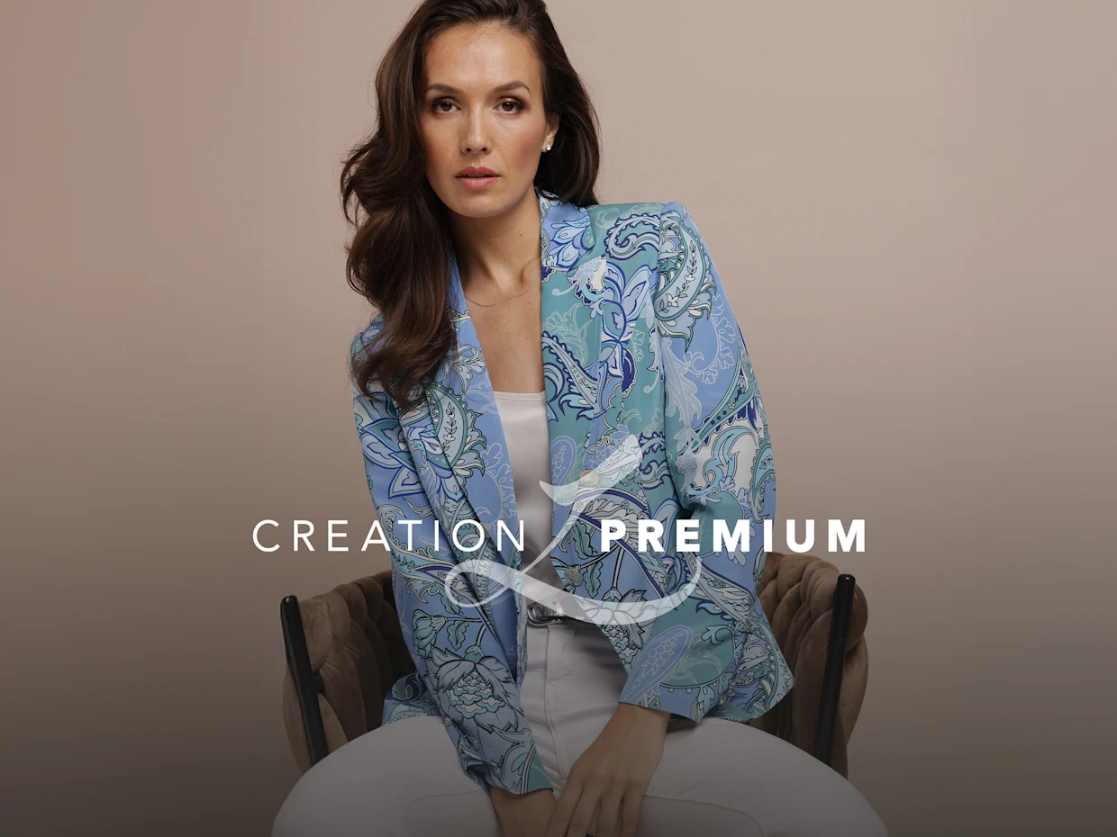 Ein Model sitzt auf einem Stuhl, trägt einen gemusterten Blazer in Blau- und Grüntönen zu weißer Hose. Text: CREATION PREMIUM.