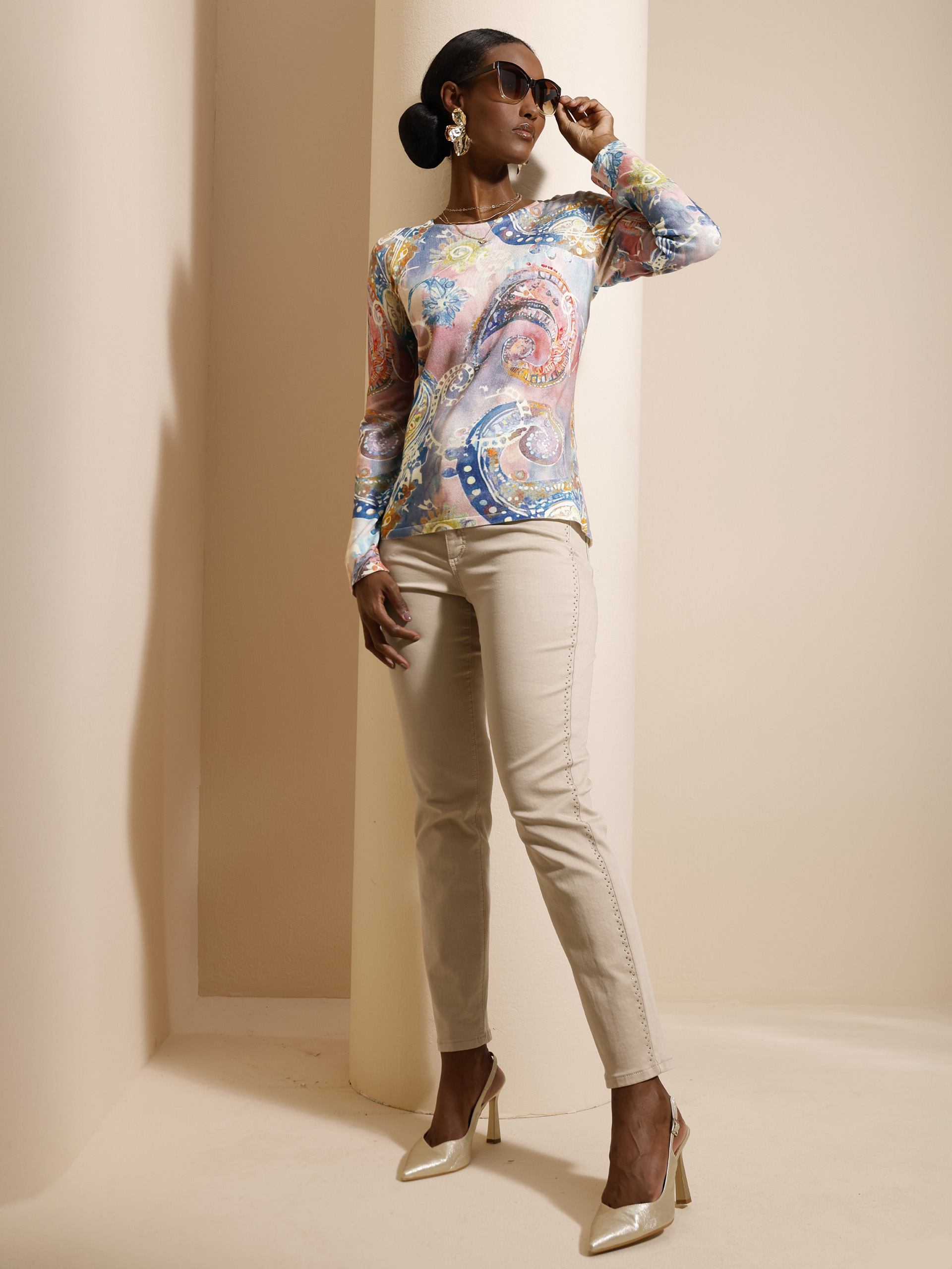 Ein Model steht vor beigem Hintergrund, trägt eine gemusterte, bunte Langarmbluse, eine beigefarbene Hose und cremefarbene High Heels.