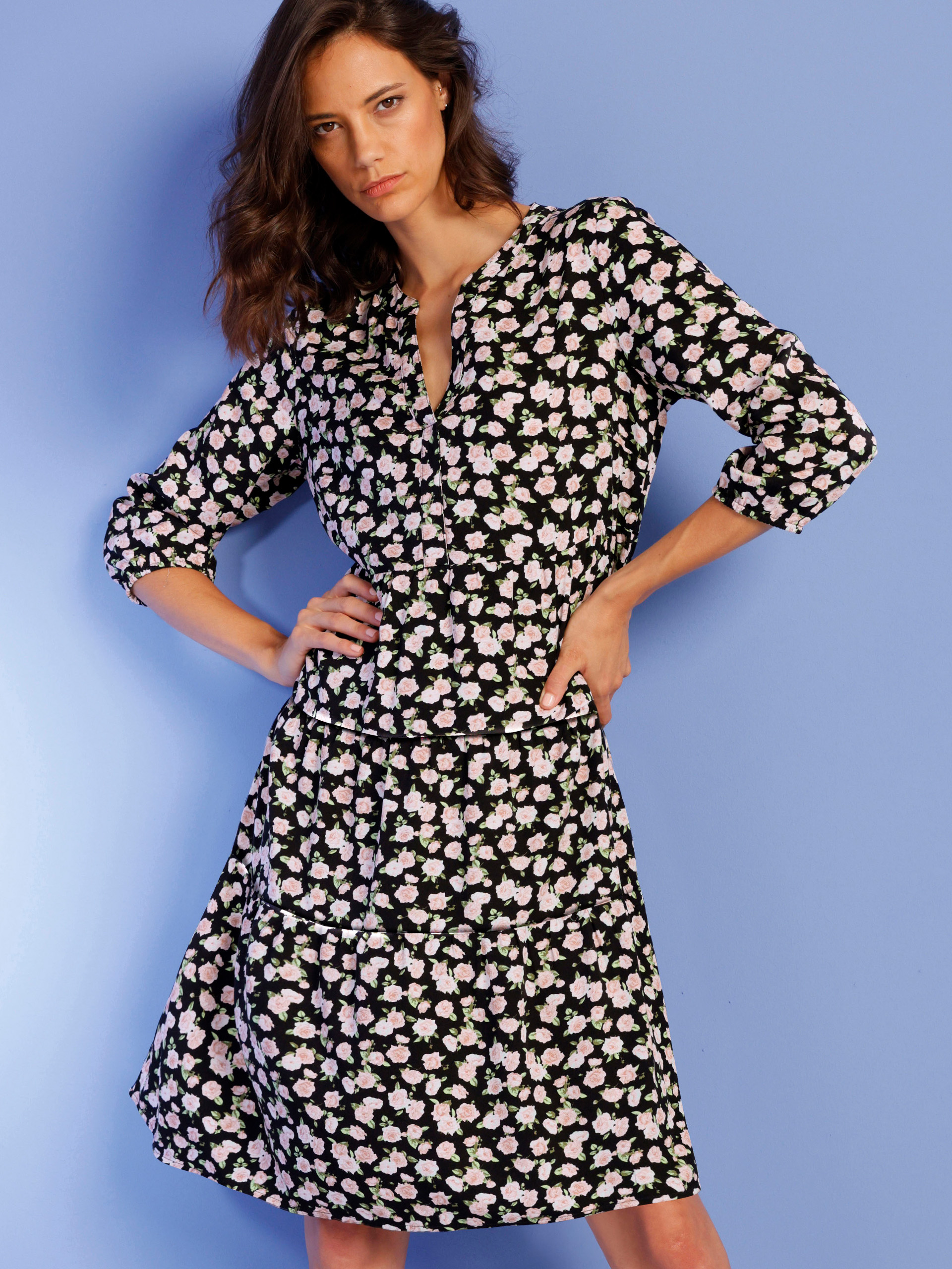 Ein Model trägt ein schwarzes Kleid mit weiß-rosafarbenem Blumenmuster und langen Ärmeln vor einem hellblauen Hintergrund.