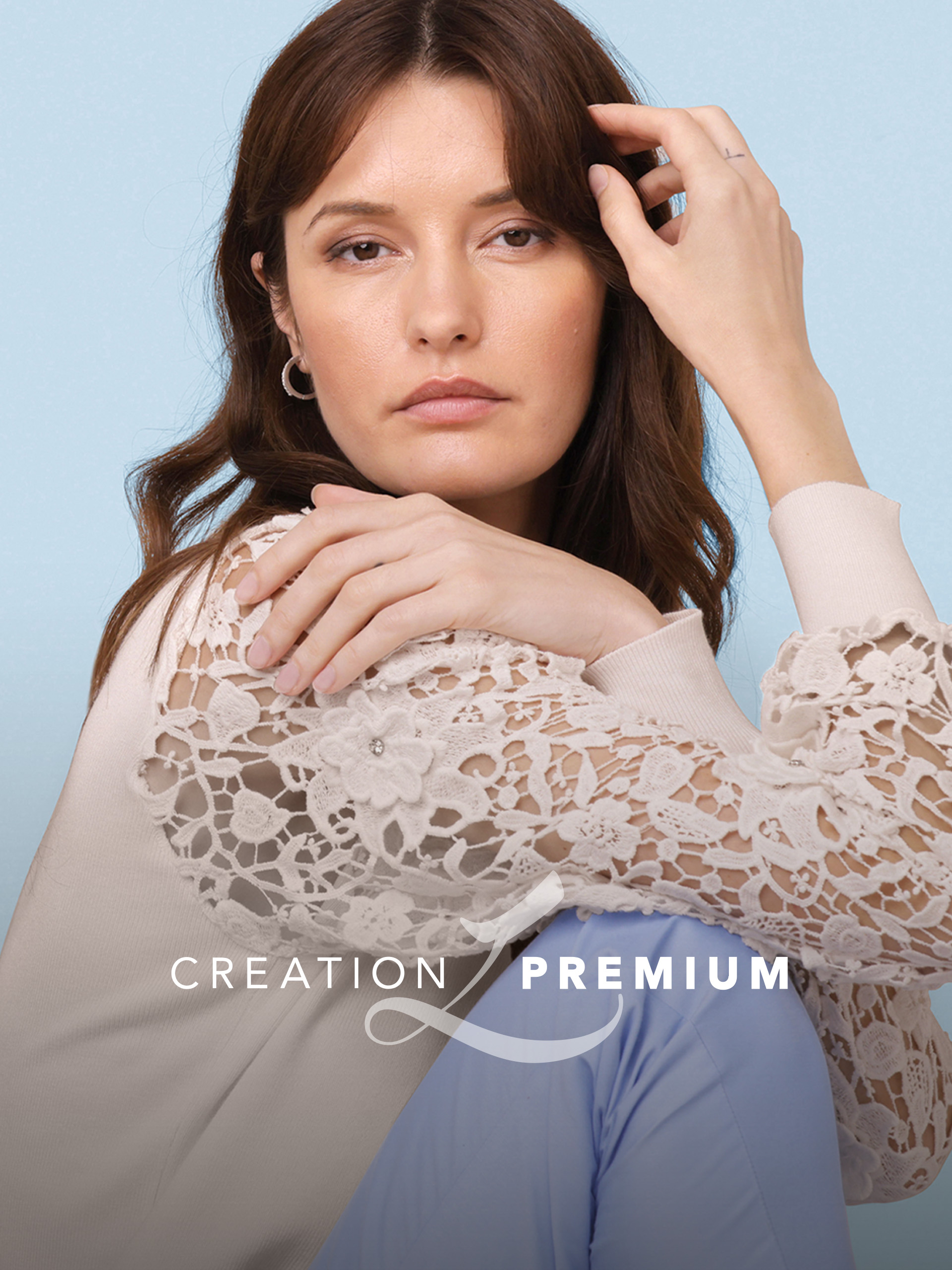 Ein Model sitzt vor hellblauem Hintergrund. Es trägt eine cremefarbene Bluse mit Spitzenärmeln und eine hellblaue Hose. Text: 'CREATION PREMIUM'.
