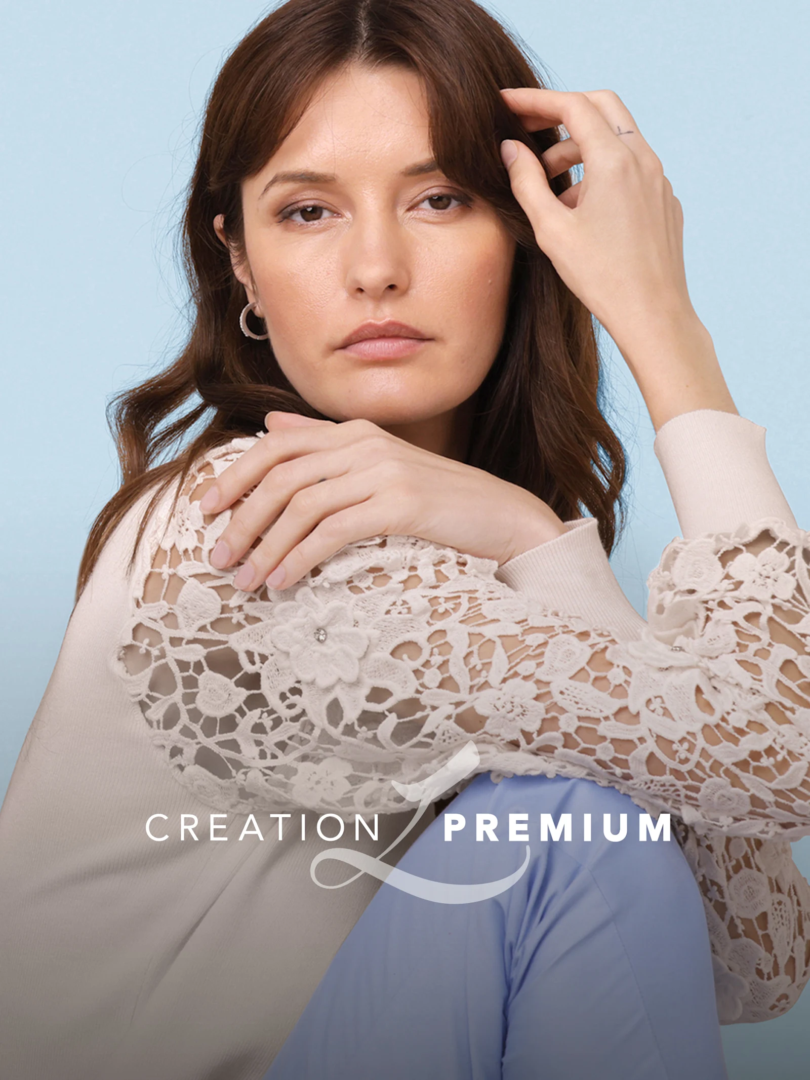 Ein Model sitzt vor hellblauem Hintergrund. Es trägt eine cremefarbene Bluse mit Spitzenärmeln und eine hellblaue Hose. Text: 'CREATION PREMIUM'.