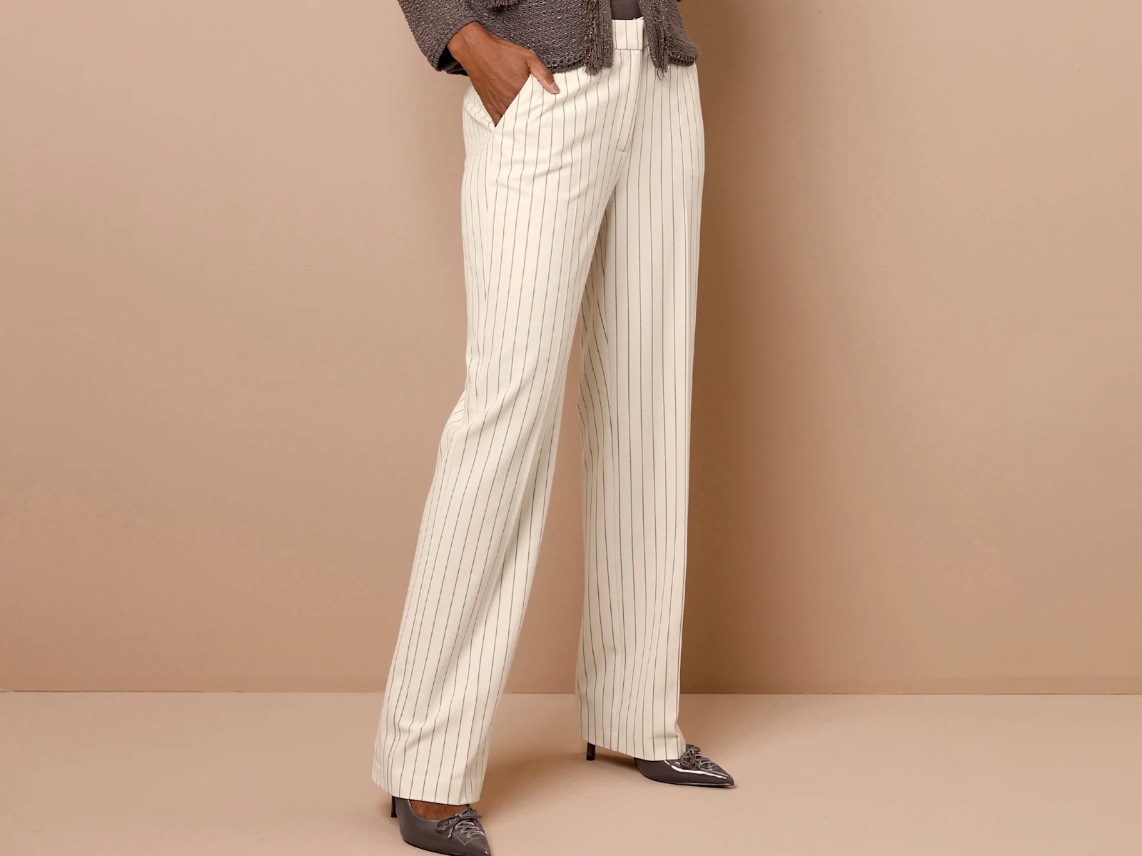 Ein Model trägt eine cremefarbene, weit geschnittene Hose mit feinen Längsstreifen und spitze, dunkle Schuhe. Der Hintergrund ist beige.