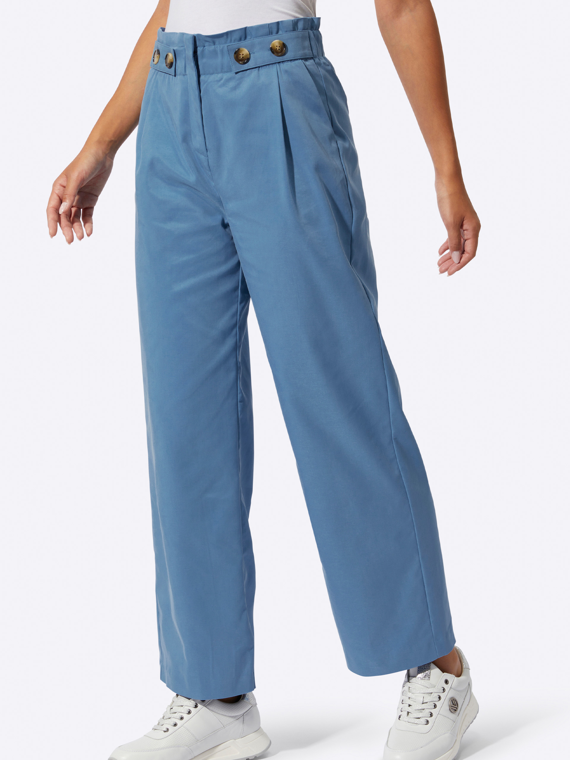 Ein Model trägt eine weite, blaue Hose mit Bundfalten und auffälligen Knöpfen sowie weiße Sneaker. Der Hintergrund ist hell.
