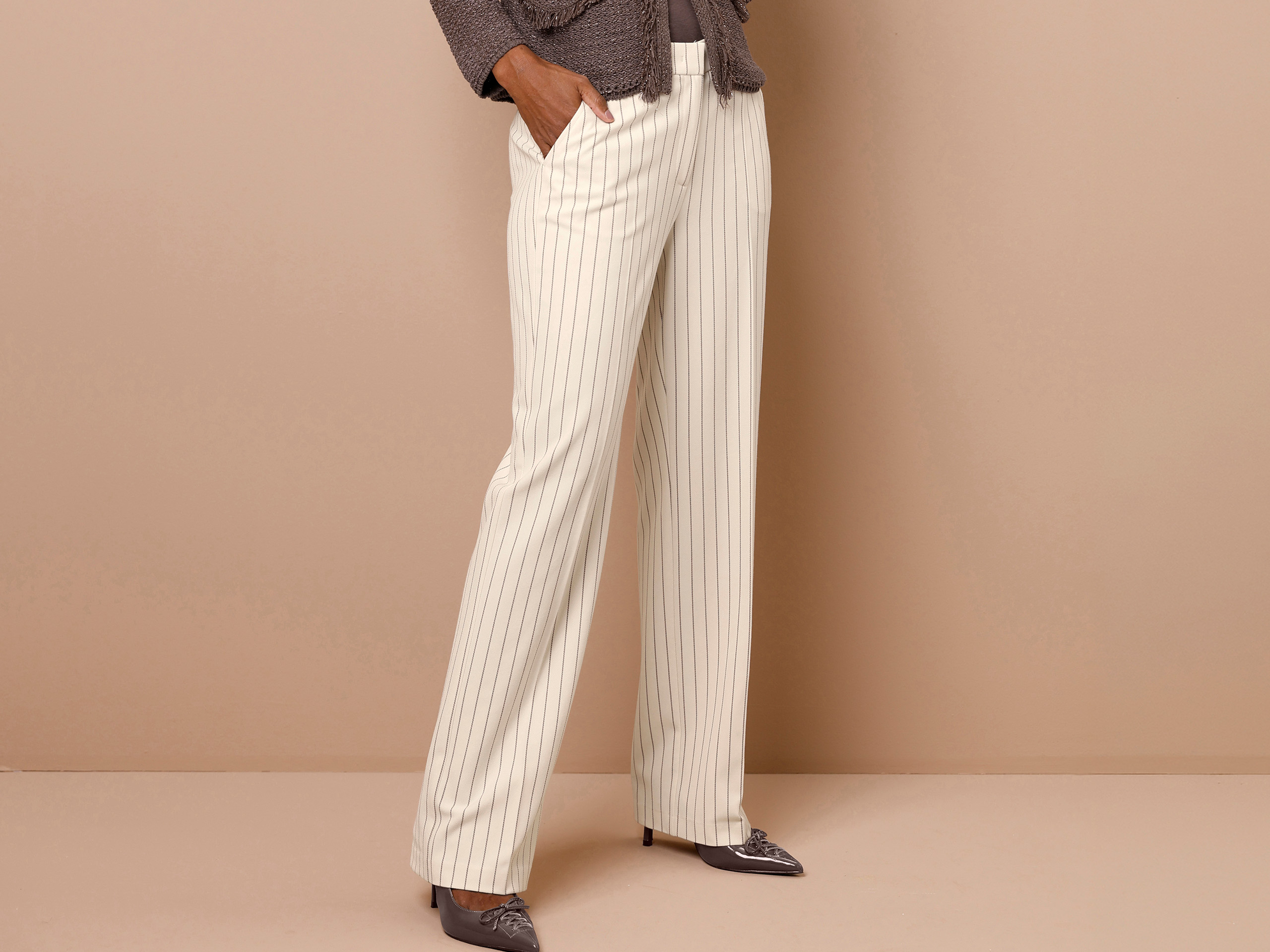 Ein Model' trägt eine cremefarbene Hose mit feinen Längsstreifen, braune Jacke, spitze graue Schuhe, Hände in den Hosentaschen.