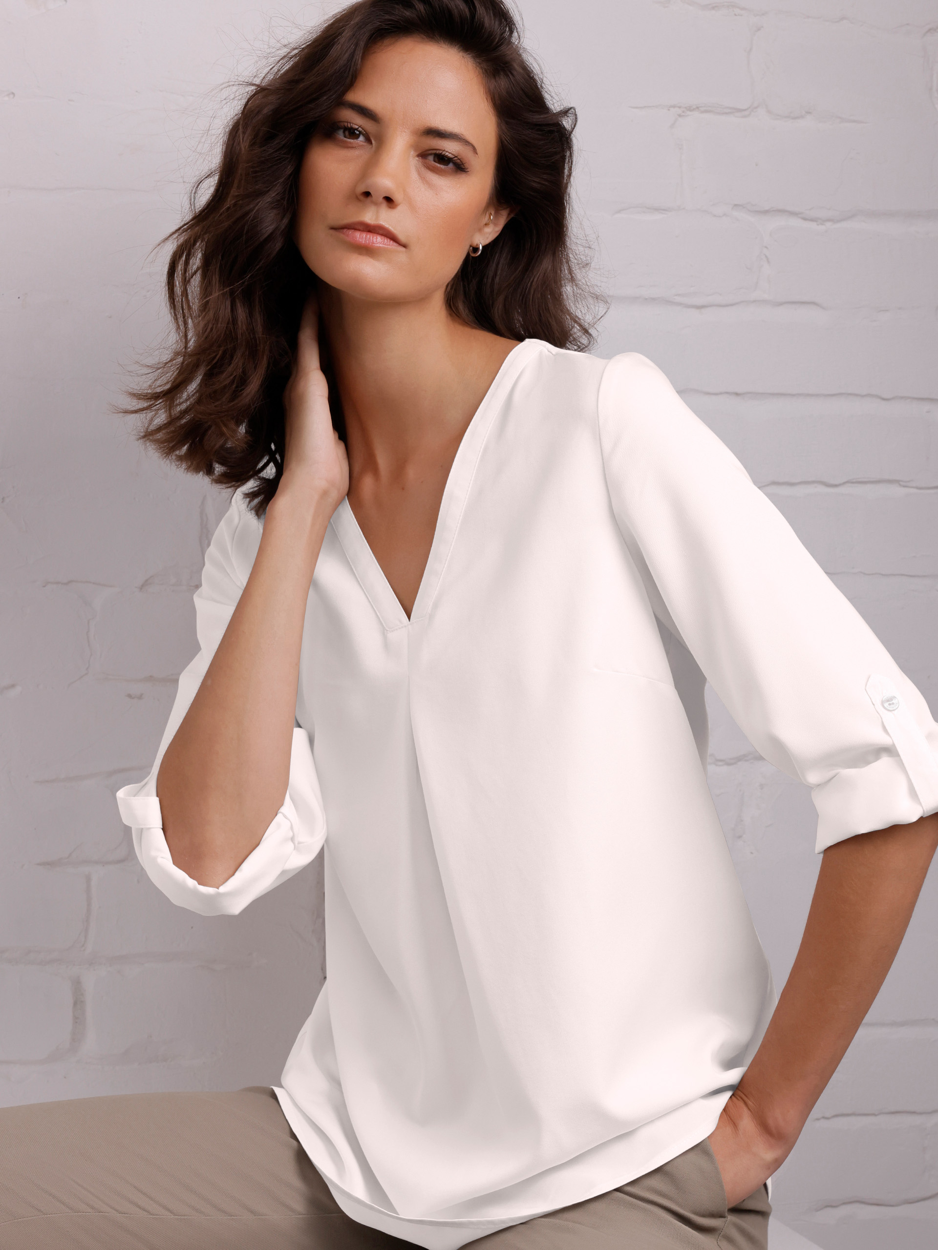 Ein Model sitzt vor einer weißen Wand und trägt eine helle Bluse mit V-Ausschnitt und Dreiviertelärmeln. Die Pose wirkt entspannt.