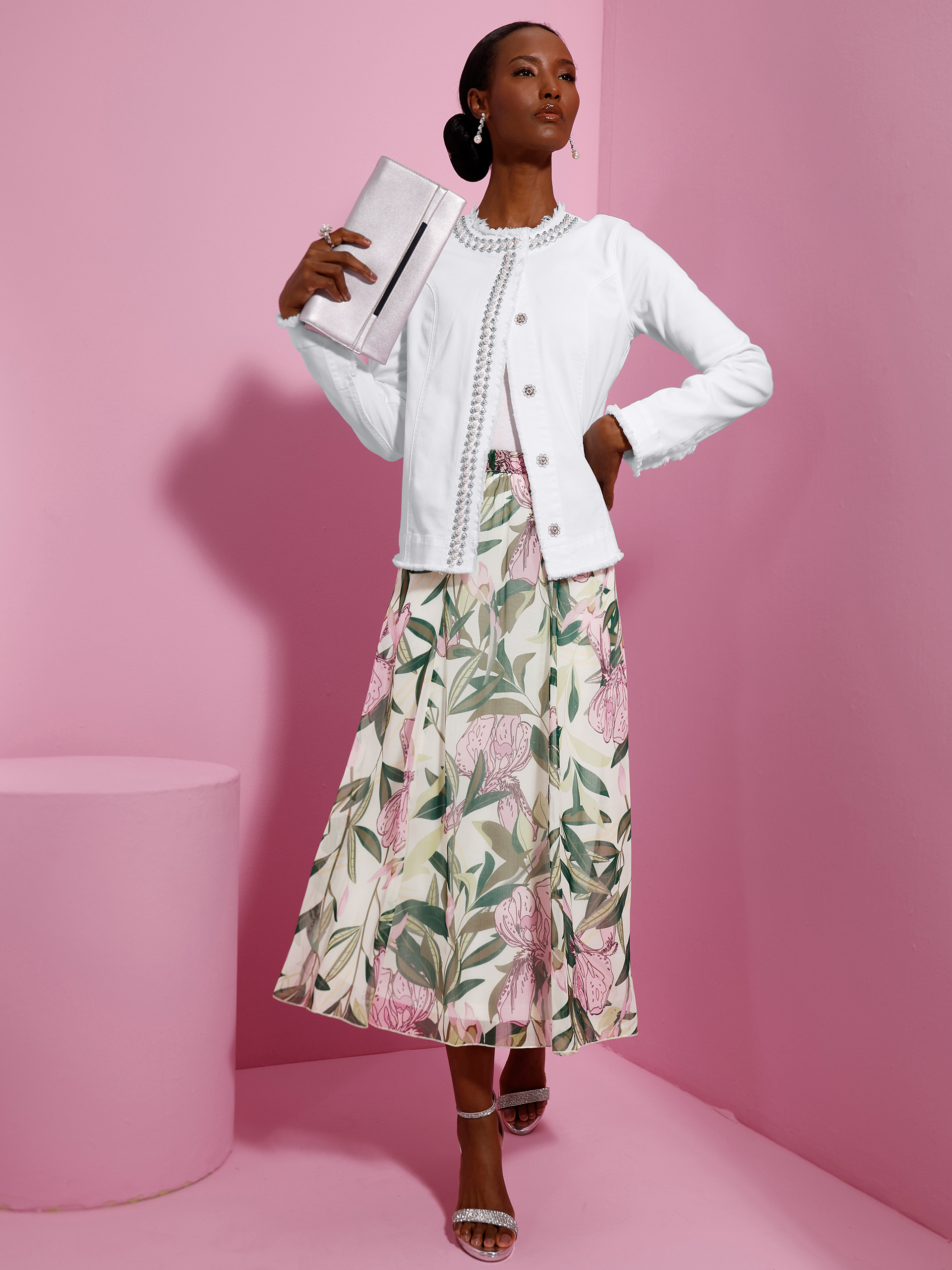 Ein Model steht vor rosa Hintergrund, trägt eine weiße Jacke, einen langen geblümten Rock in Rosa-Grün, silberne Sandaletten und hält eine Clutch.
