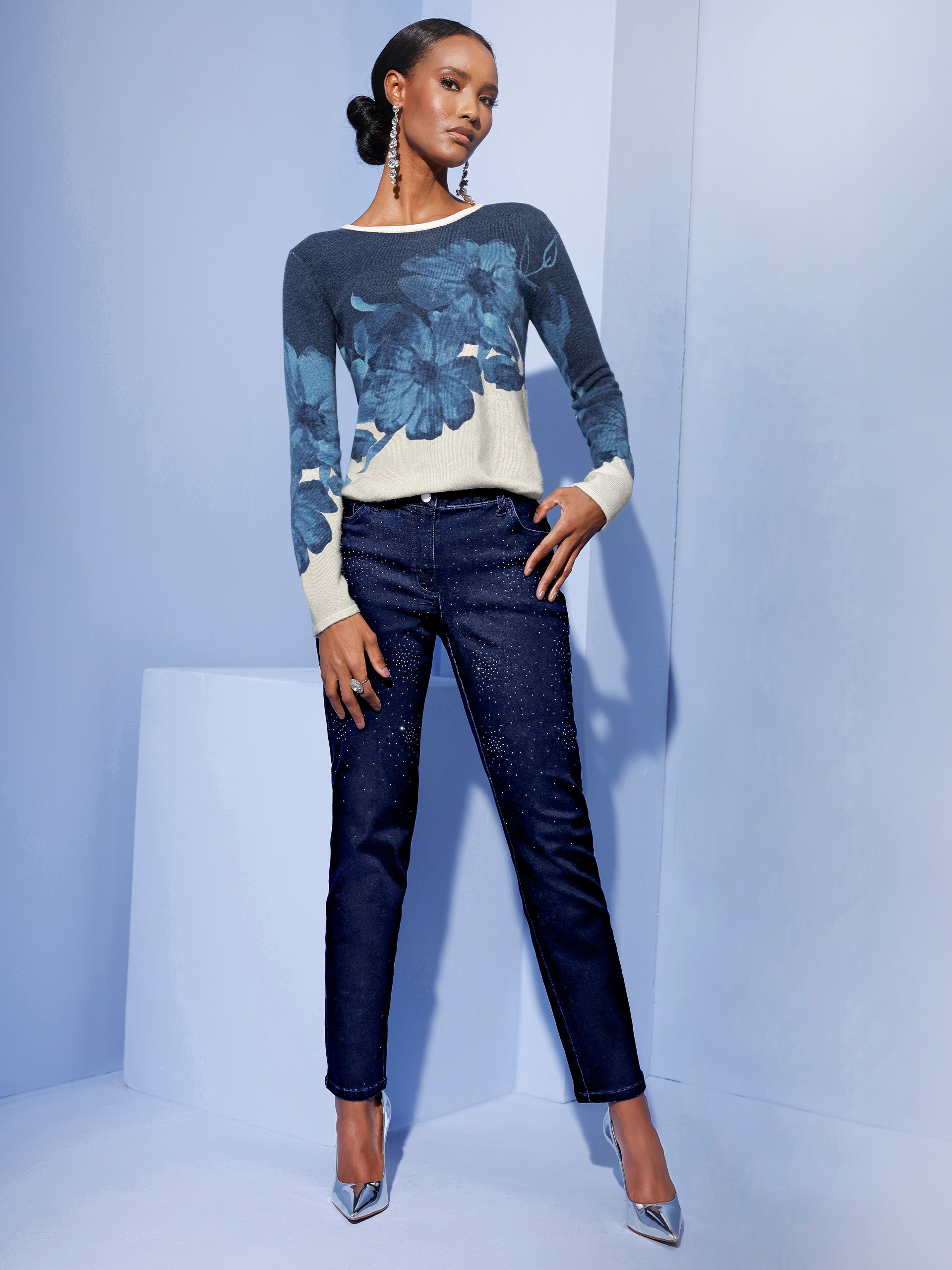 Ein Model trägt einen blauen Pullover mit floralem Muster und dunkelblaue Jeans. Die Schuhe sind silberfarben. Der Hintergrund ist hellblau.