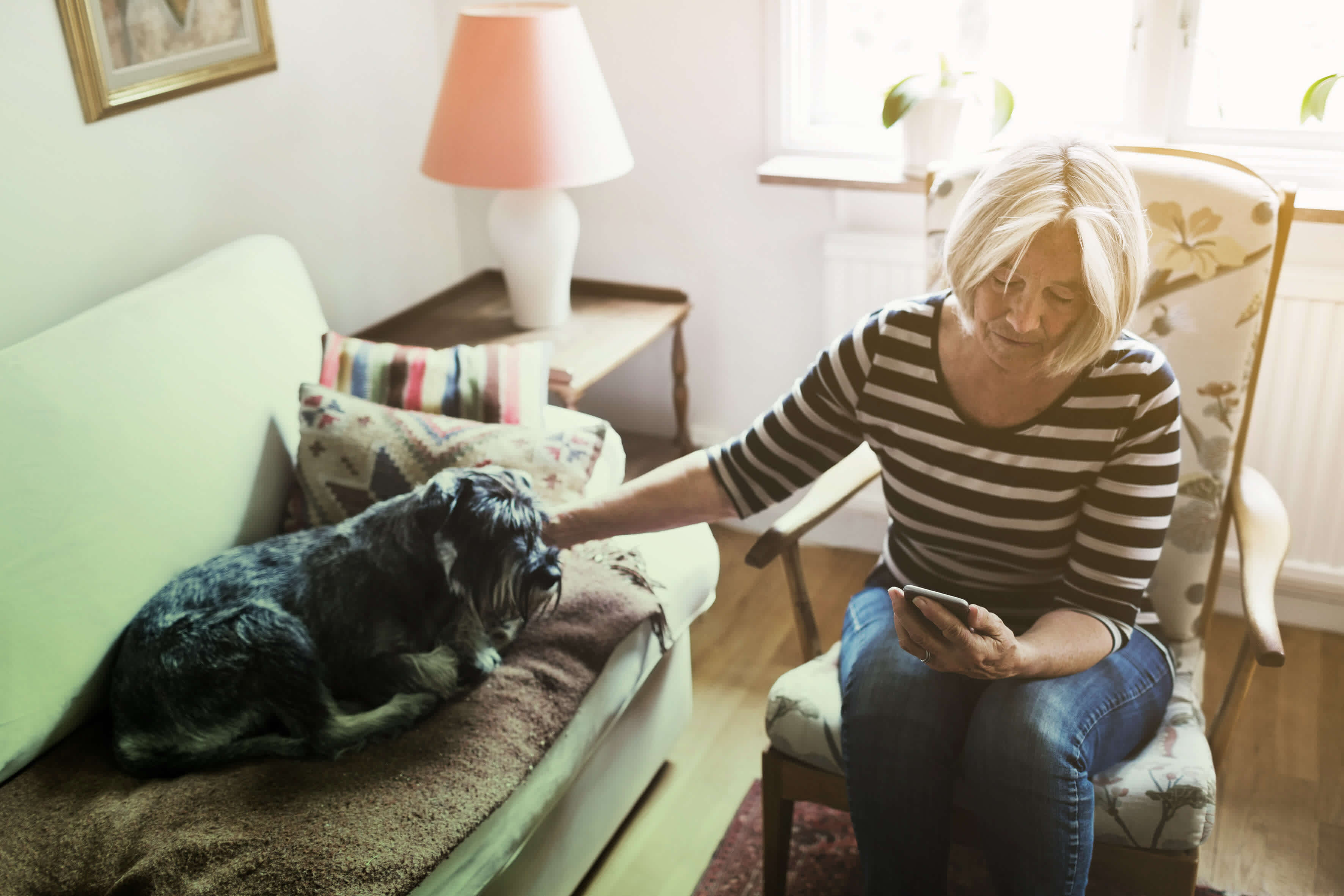 A woman is sitting at home with a dog and looking at her mobile.