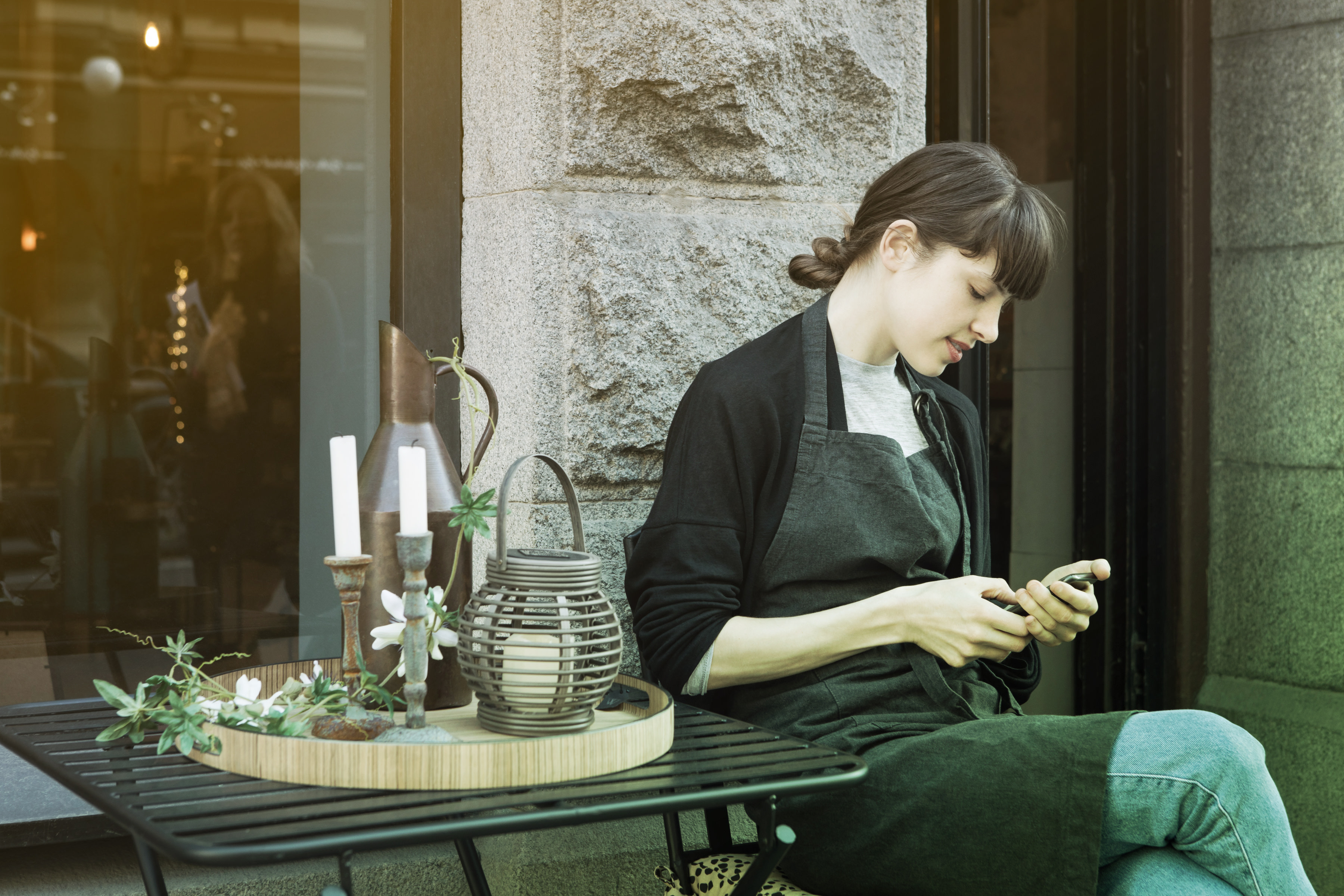 Kvinna sitter vid ett cafébord utanför en butik och kollar på sin mobiltelefon. 