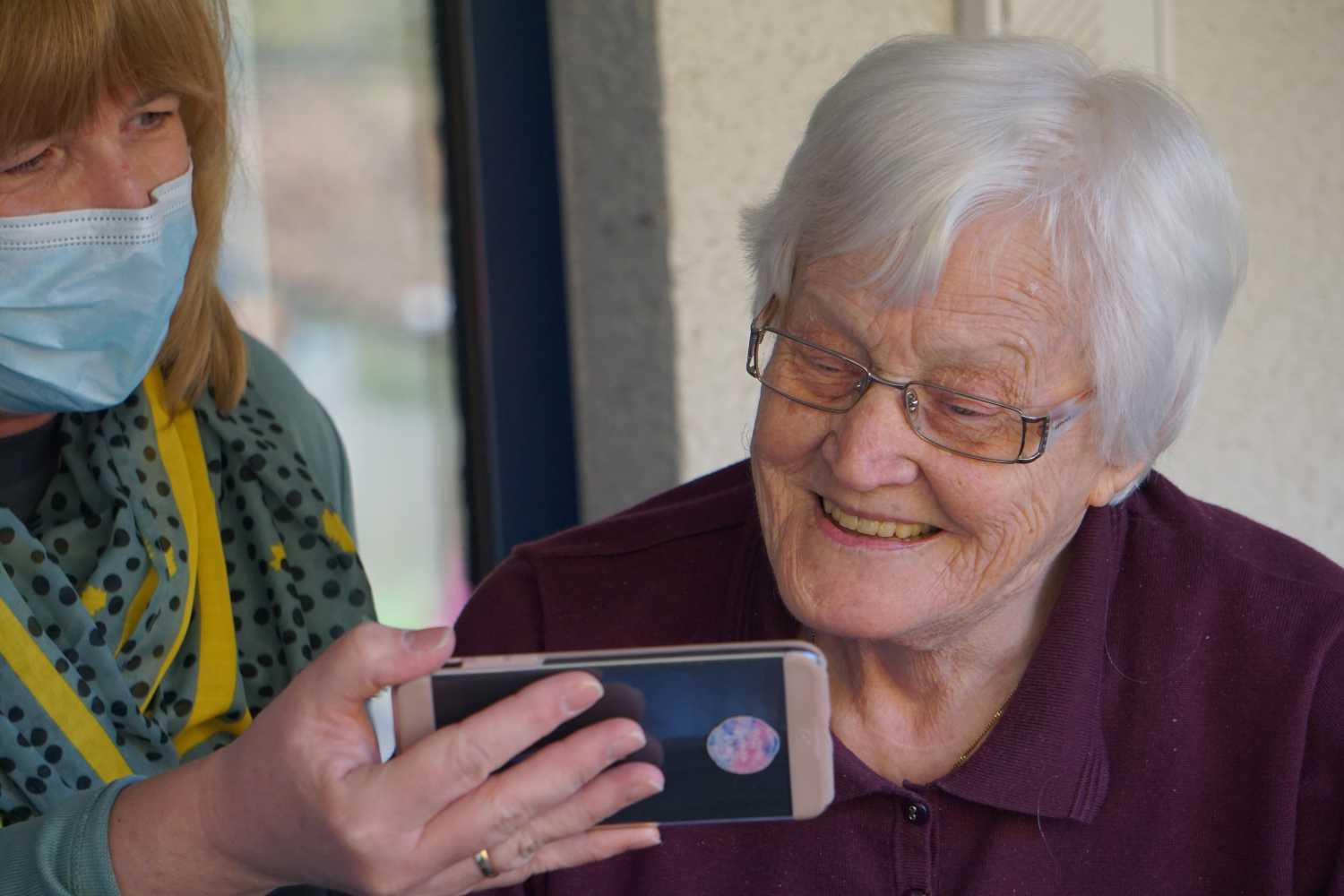 Äldre kvinna får hjälp av sköterska att titta i mobil.