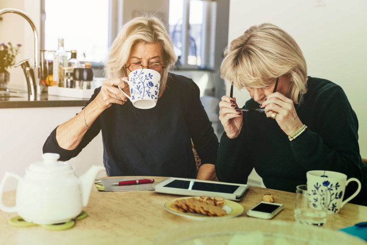 Two women are sitting at a kitchen table having coffee, while working on an iPad.