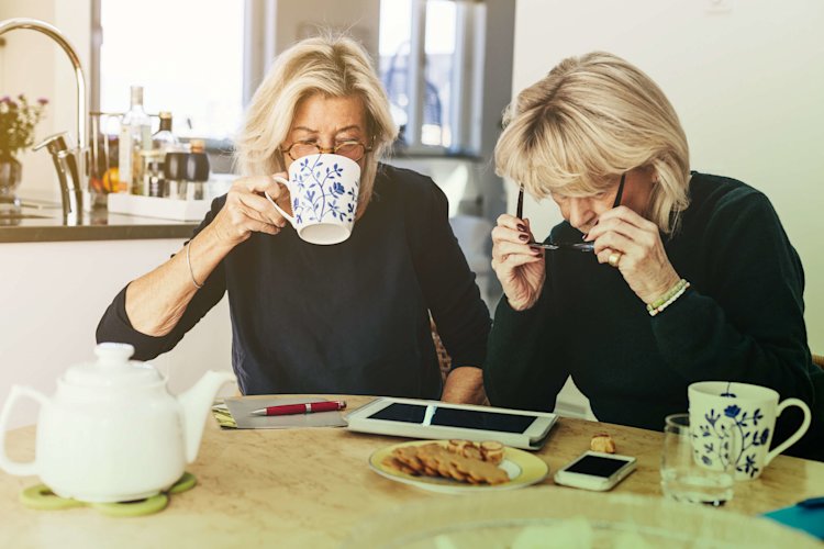 Two women are sitting at a kitchen table having coffee, while working on an iPad.