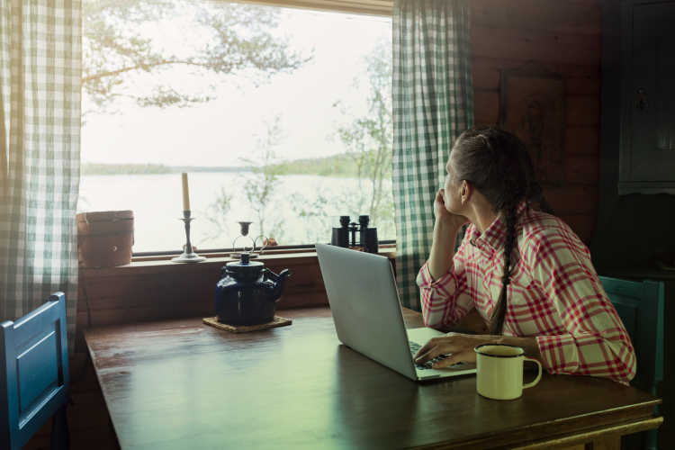 Kvinna sitter vid ett köksbord och arbetar på sin dator, men har blicken fäst ut genom fönstret. 