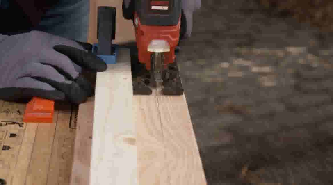 GAMMA klusser met werkhandschoenen zaagt een houten plank met een oranje decoupeerzaag, vastgeklemd met een oranje klem.