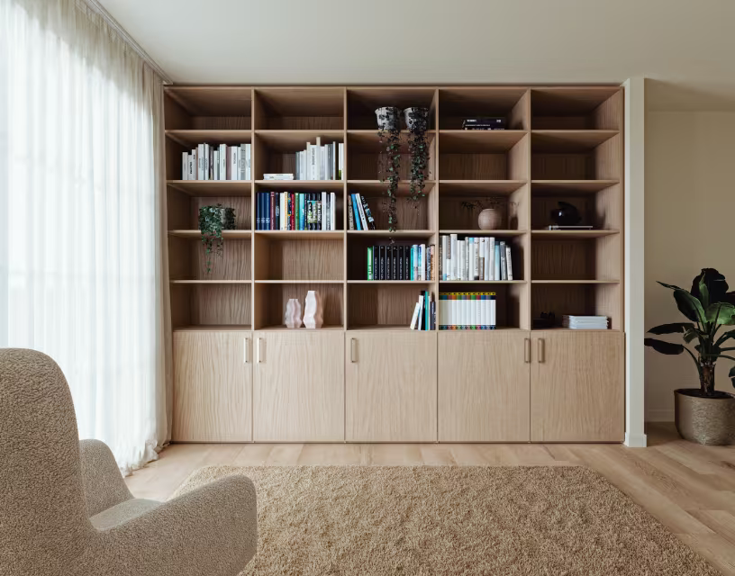 Grote houten boekenkast met open vakken en gesloten kasten, gevuld met boeken en decoratieve objecten in moderne woonkamer.