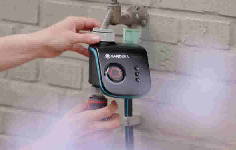 Hand installeert een zwart Gardena bewateringssysteem met rode knop aan een buitenkraan op een witte muur.