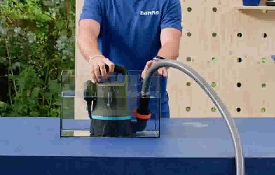 Persoon in blauw Gamma-shirt demonstreert een dompelpomp in een transparante bak op een blauwe tafel.