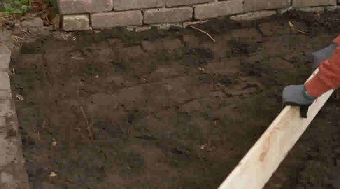 Gehandschoende hand houdt houten plank vast boven omgewoelde donkere aarde naast een stenen muurtje in een tuin.