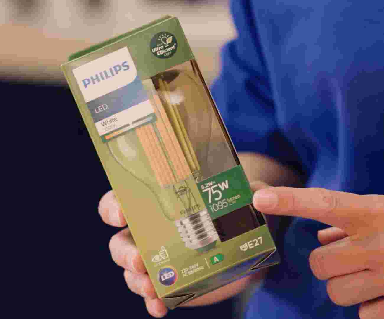 Iemand in een blauwe GAMMA trui houdt een verpakking van een Philips LED-lamp vast en wijst daarbij naar het aantal lumen en wattage. 