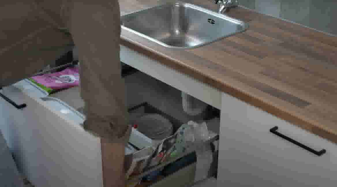 Persoon opent een keukenkastje onder een houten aanrecht met roestvrijstalen spoelbak, gevuld met schoonmaakartikelen.