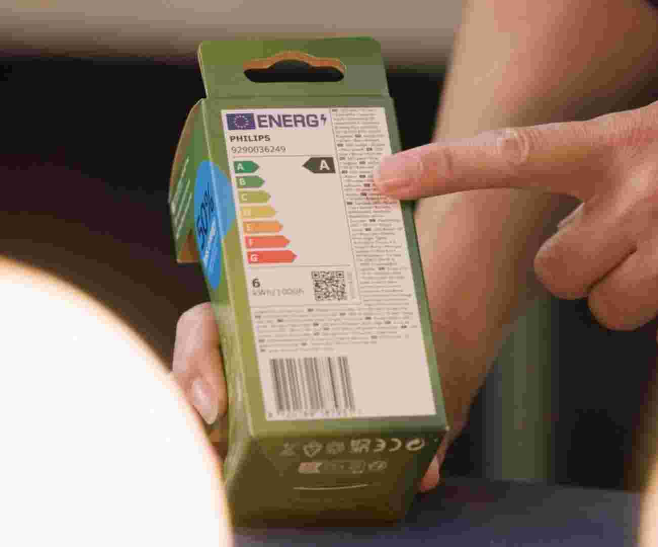 Iemand houdt een verpakking van een Philips LED-lamp vast en wijst daarbij naar het energielabel Deze loopt van A tot en met G. De LED lamp in de verpakking heeft label A. 