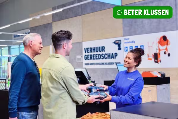 GAMMA medewerker in blauwe trui overhandigt gereedschap aan klant bij verhuurbalie in de bouwmarkt met "Beter Klussen" logo en "Verhuur Gereedschap" bord op de achtergrond.