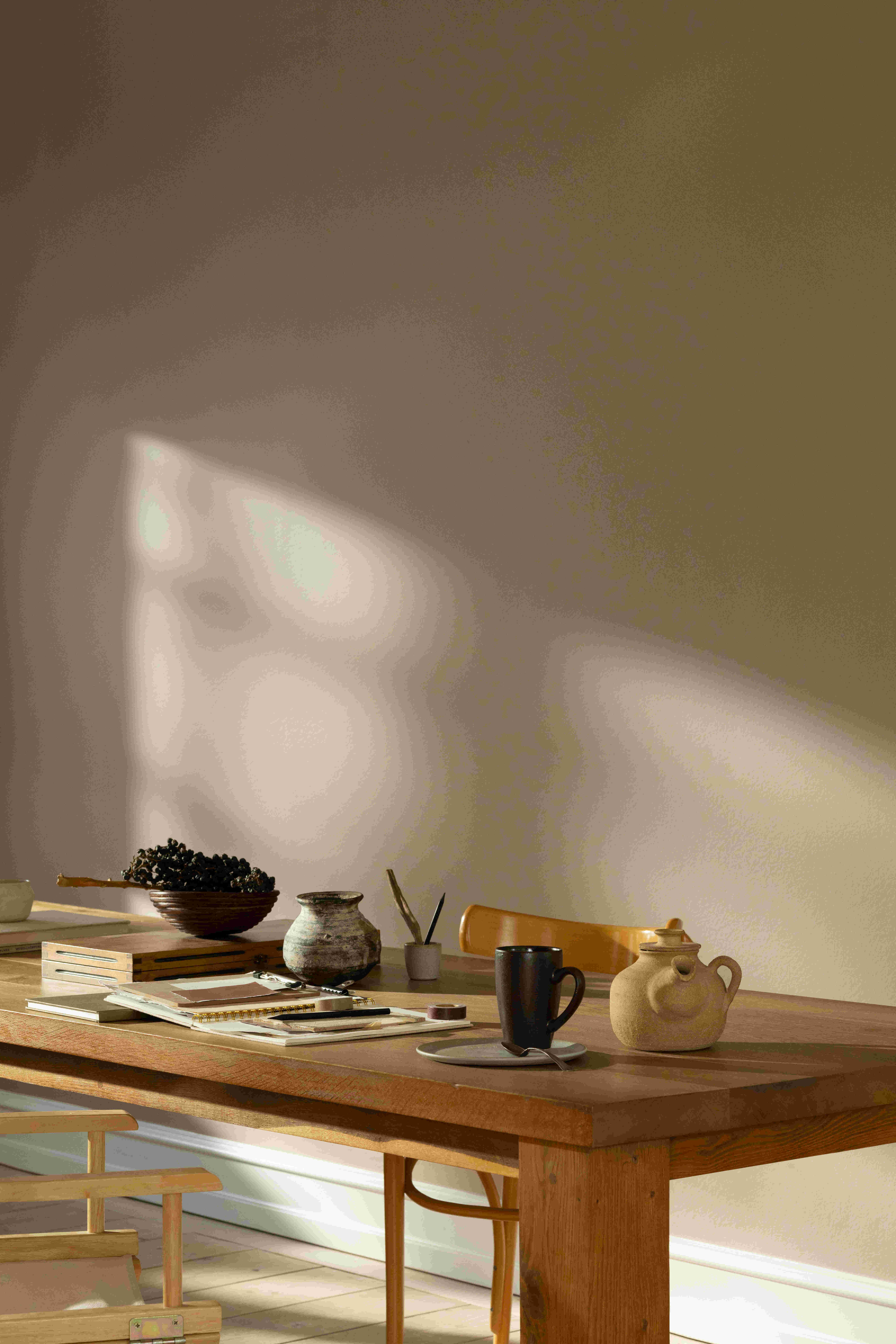 Houten tafel met boeken, koffiemok, theepot en decoratieve objecten in warm zonlicht met Country Soil kleur op de muur