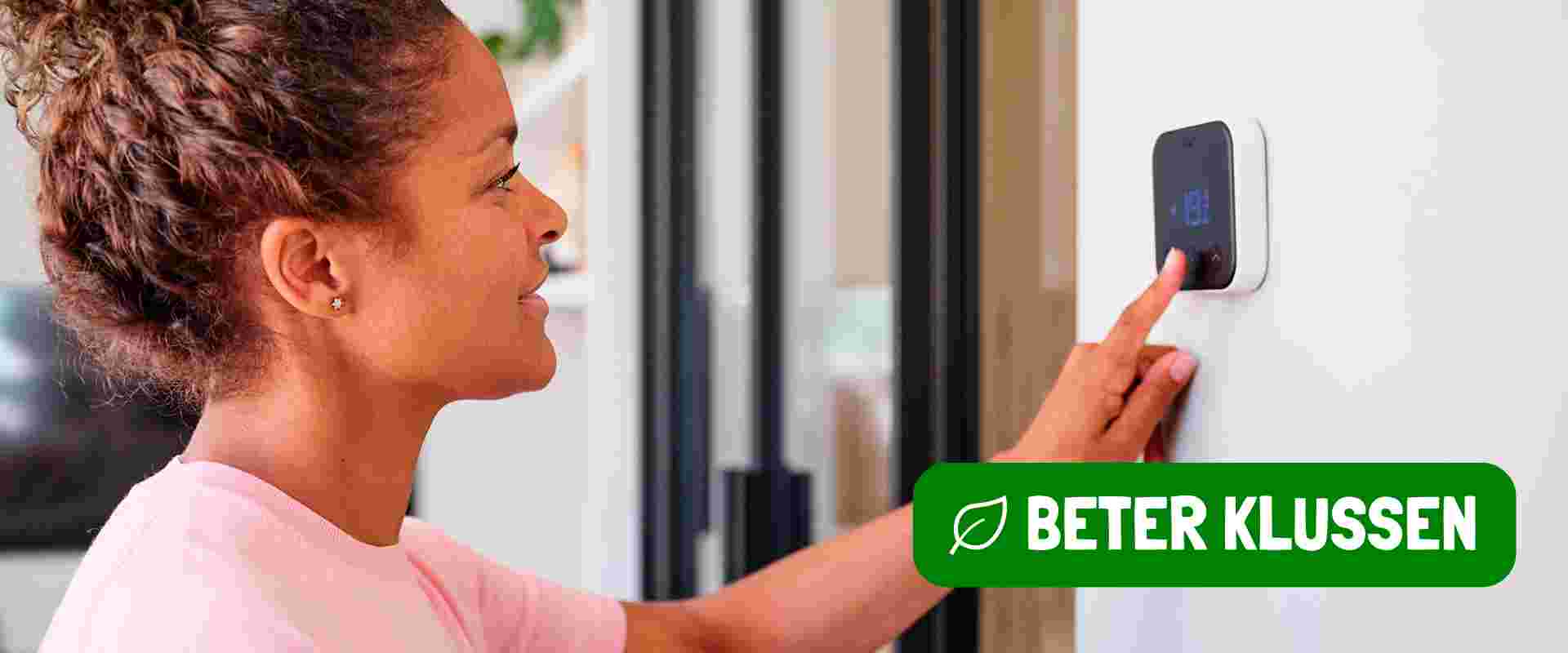 Een vrouw in roze shirt bedient een slimme thermostaat aan de muur, met "BETER KLUSSEN" logo in groene balk onderaan.