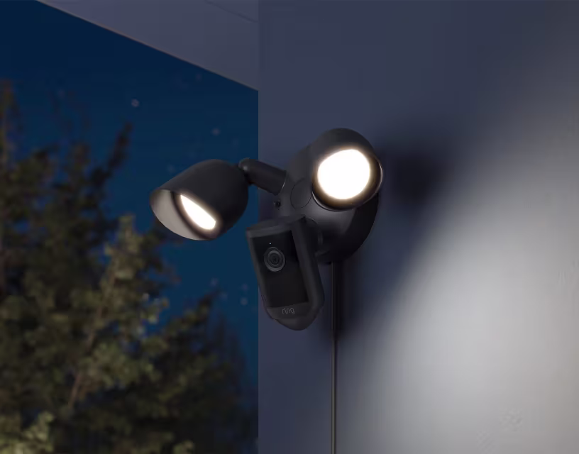 Zwarte buitenlamp met twee verlichte LED-spots en geïntegreerde beveiligingscamera op witte muur bij schemering.