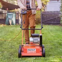 Man in beige jas bedient een gehuurde verticuteermachine op een groen gazon in een zonnige achtertuin.