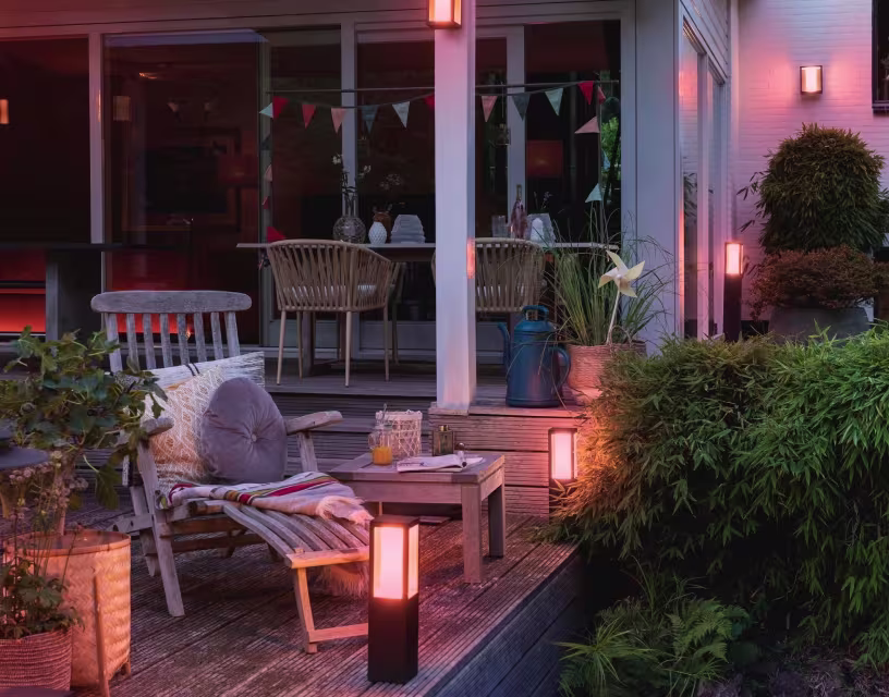 Gezellig verlicht terras bij schemering met houten tuinmeubelen, lantaarns, planten en kleurrijke vlaggetjes aan het huis.