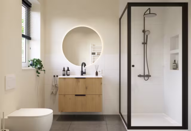 Moderne badkamer met ronde verlichte spiegel, houten wastafelmeubel, zwarte accenten en glazen douchecabine met zwart frame.