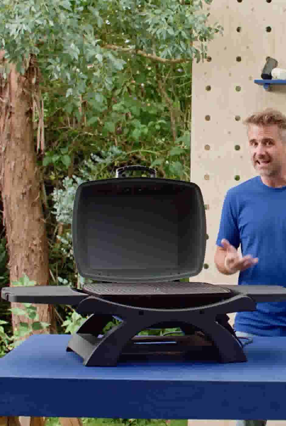 Man in blauw shirt staat naast een zwarte elektrische barbecue op een blauwe tafel, met groene planten op de achtergrond.