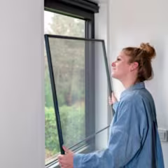 Persoon in blauw overhemd installeert een hor voor een raam met uitzicht op groen landschap.
