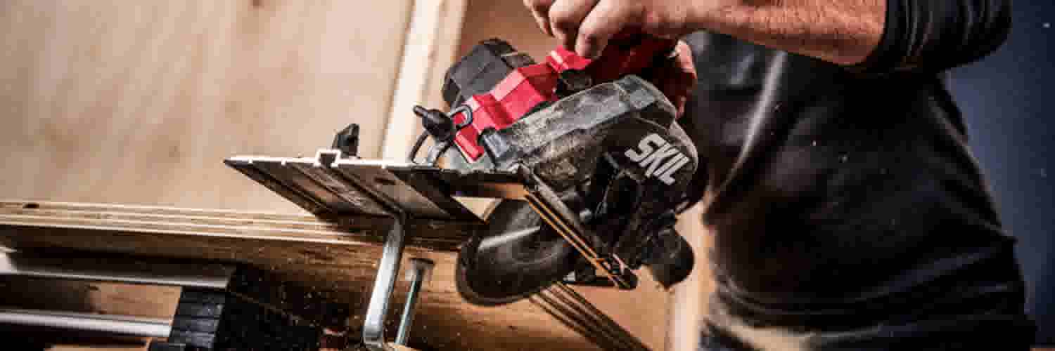 Handen van een vakman die een rood-zwarte SKIL cirkelzaag gebruikt om hout te zagen op een werkbank.