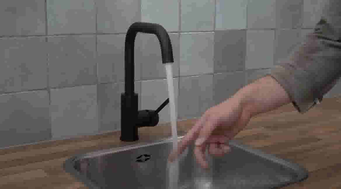 Hand reikt naar stromend water uit een zwarte kraan boven een roestvrijstalen gootsteen in een keuken met blauwe tegels.