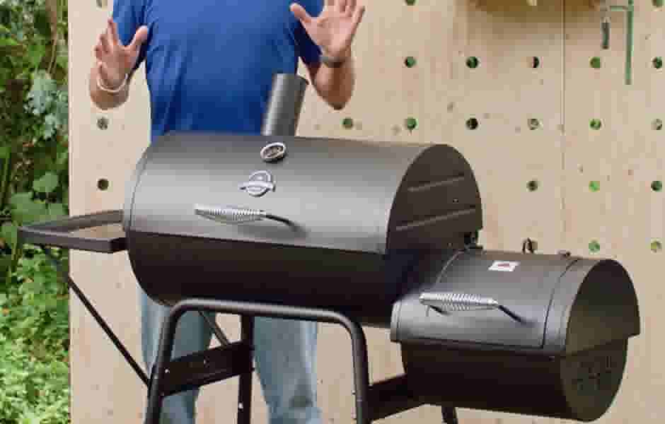 Persoon in blauw shirt staat achter een zwarte barbecue met rookoven, met opgeheven handen voor een houten wand.