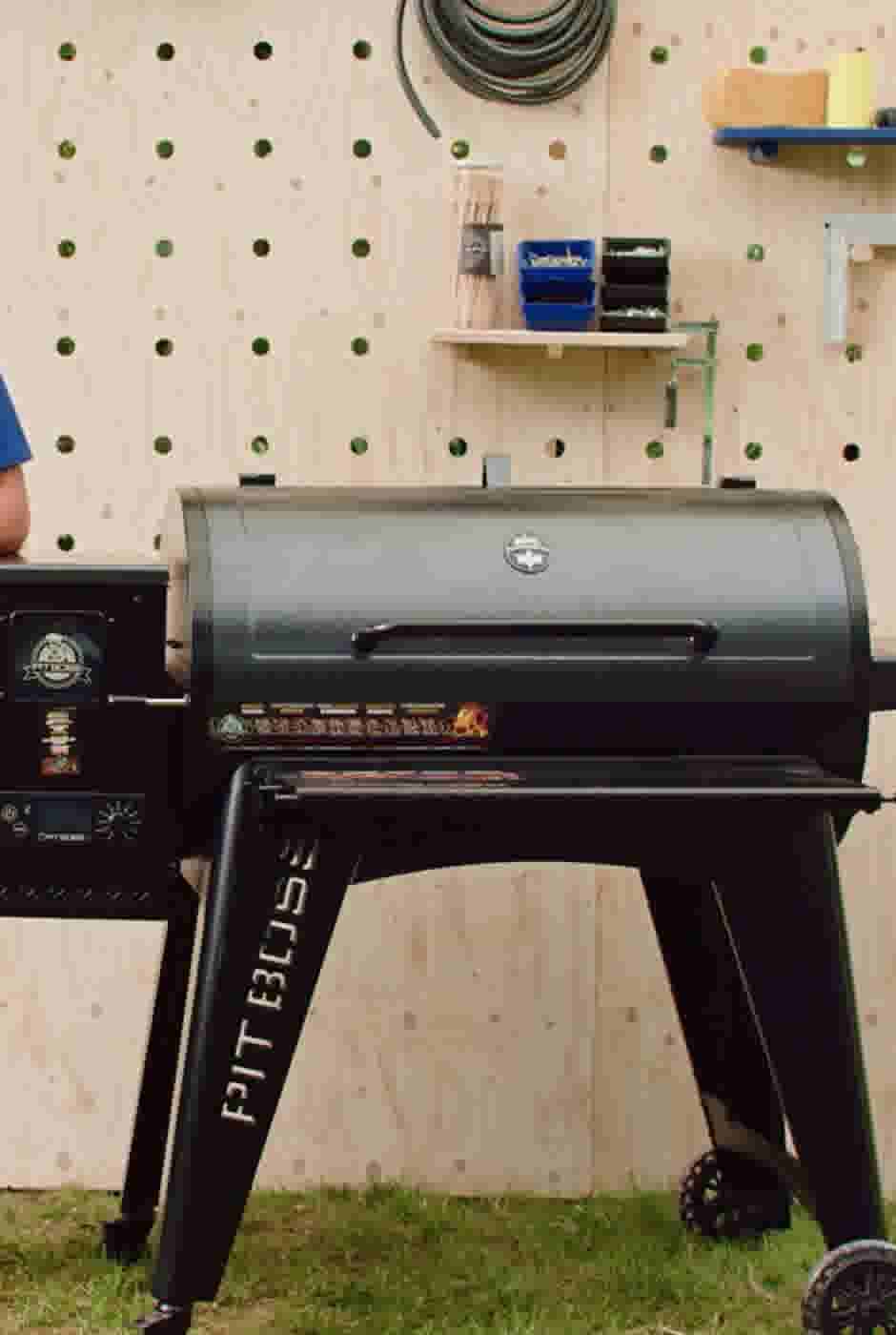 Zwarte Pit Boss barbecue op gras voor een houten wand met gereedschapsplank en opgehangen tuinslang.