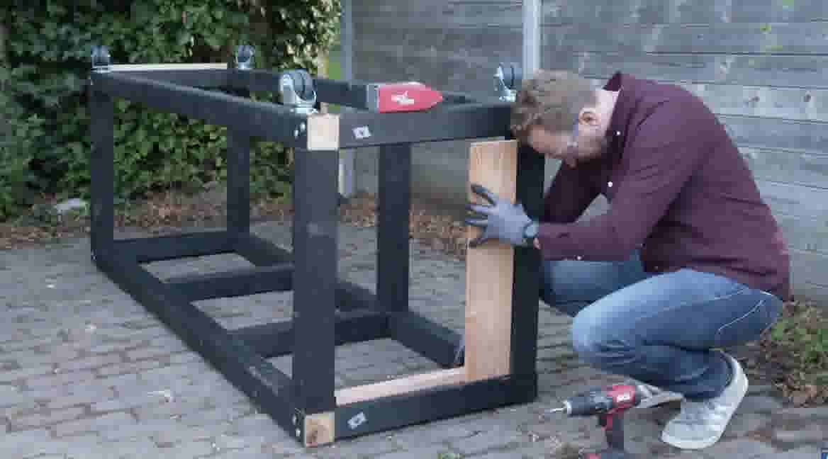 GAMMA klusser bevestigt een houten plank aan een zwart metalen frame in een tuin, met een boormachine op de grond.