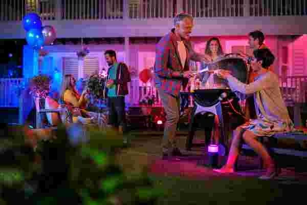 Avondfeest in de tuin met een barbecue en kleurrijke Philips Hue verlichting. Je ziet mensen genieten van eten en gesprekken voeren.