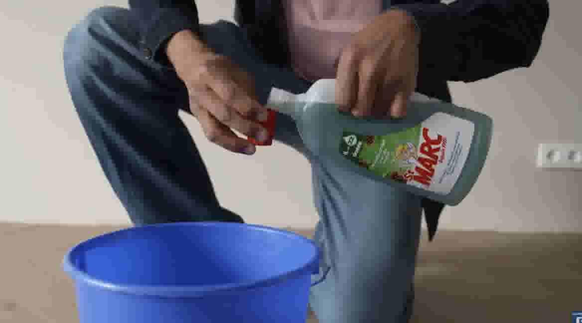 GAMMA klusser giet Marc schoonmaakmiddel in een blauwe emmer, zittend op de vloer in donkere kleding.