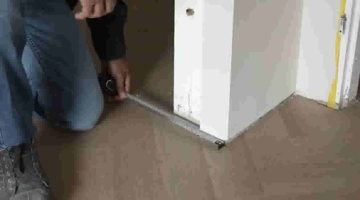 GAMMA klusser in spijkerbroek meet met rolmaat de afstand bij een deuropening op een houten vloer.